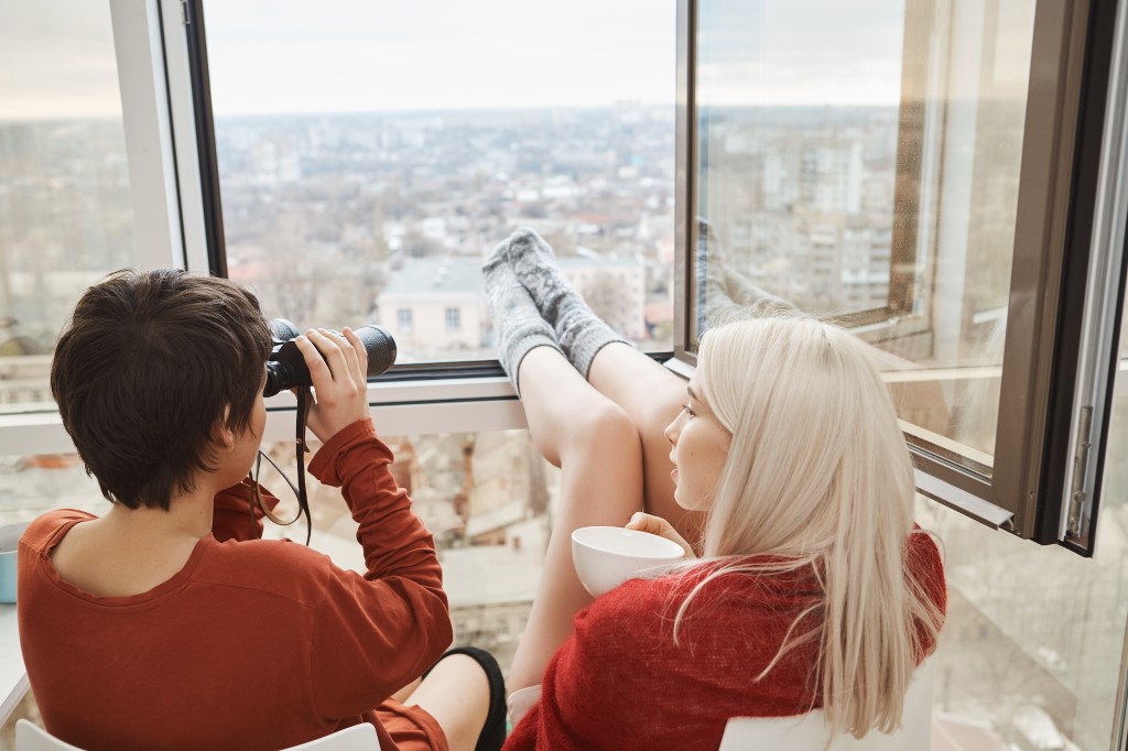 back portrait hot attractive women sitting balcony with legs leaned window using binocular drinking coffee womans fool around spy after their neighbors enjoy scenery their town